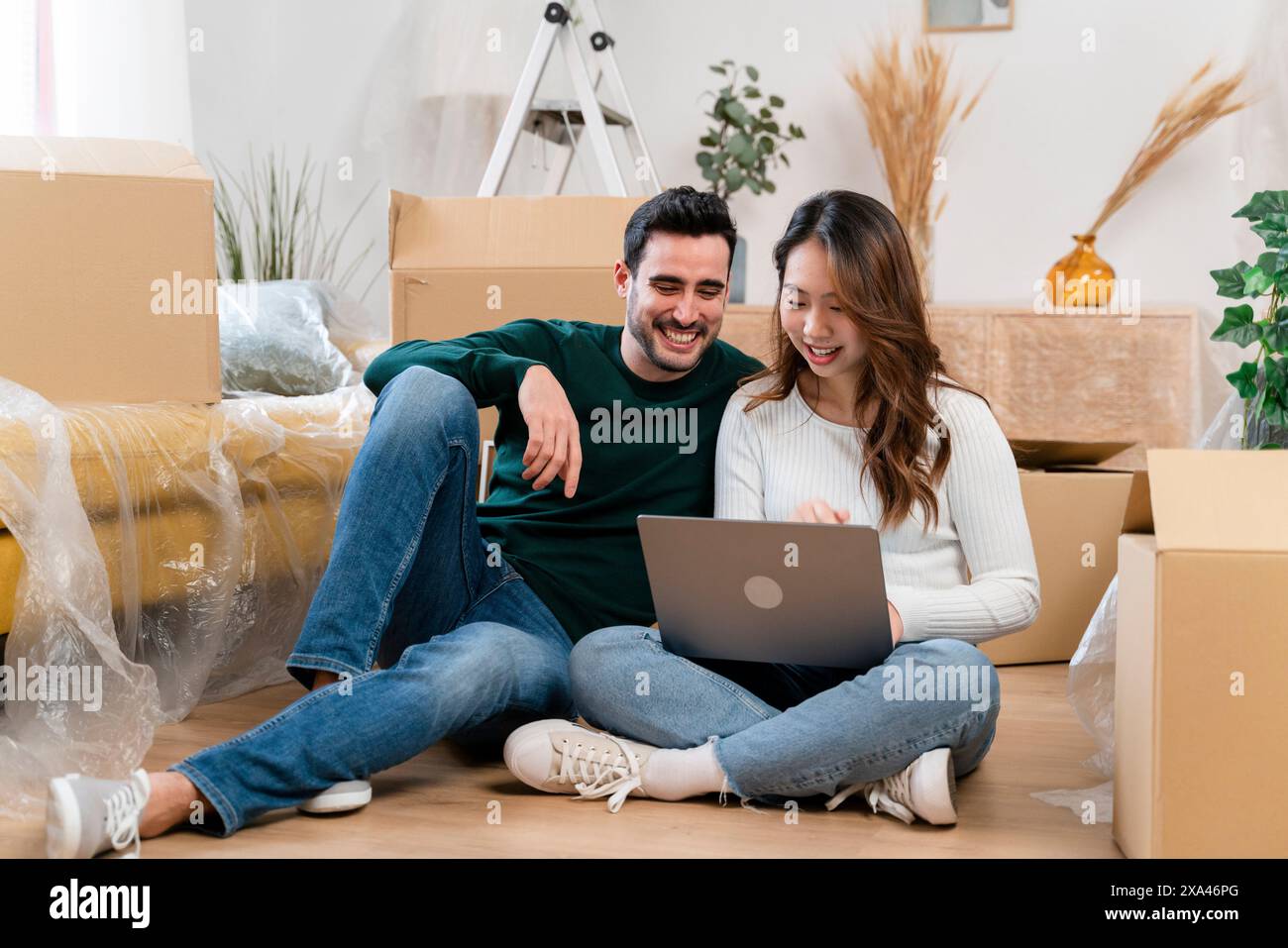 Couple smiling while using a laptop amidst moving boxes. Stock Photo