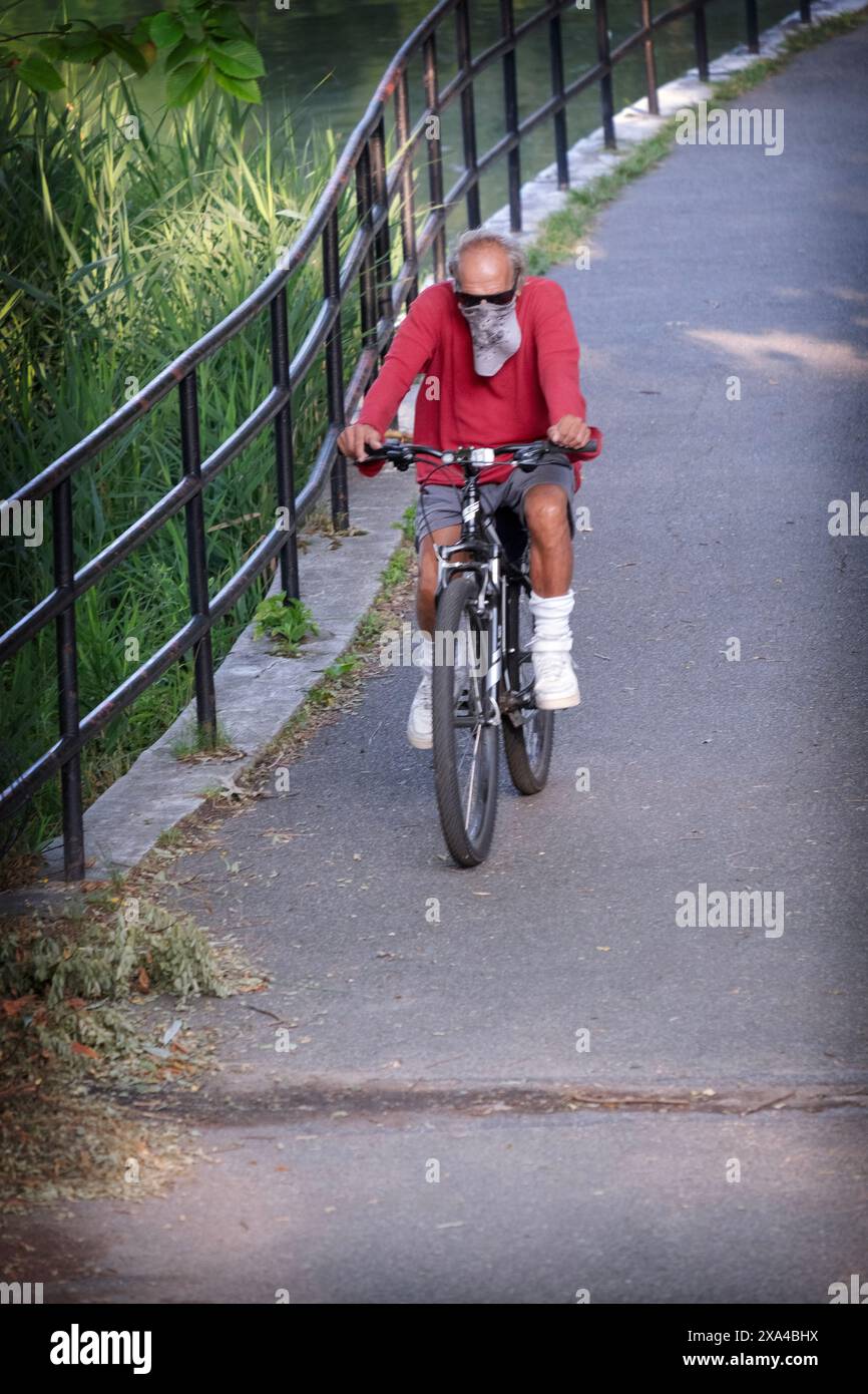 An older man wearing a bandana rides his bike on a path inear the lake in Kissena park in Flushing, Queens, New York City. Stock Photo