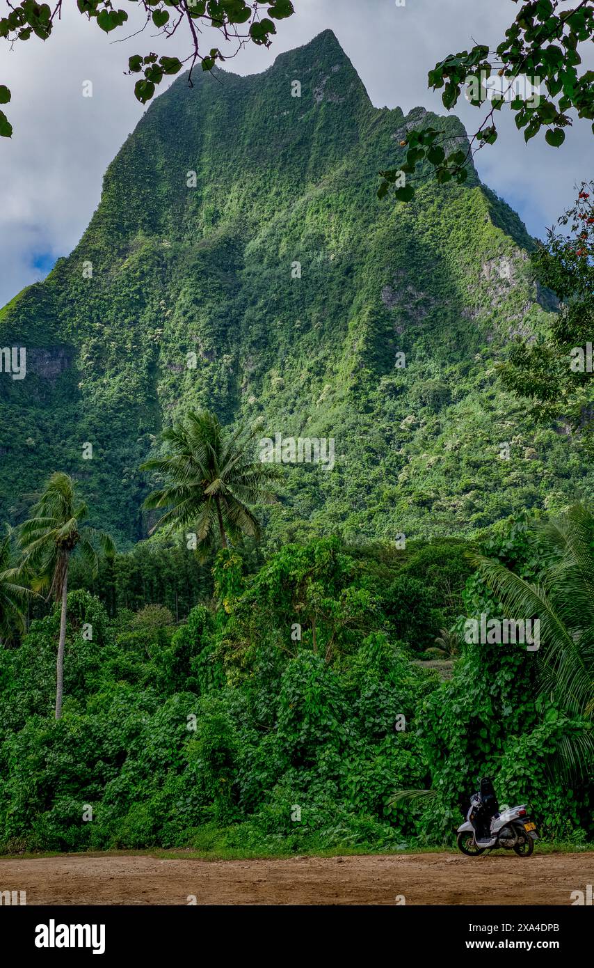 A motorcycle is parked on a dirt path with lush tropical vegetation and towering green mountain in the background under a cloudy sky. Stock Photo