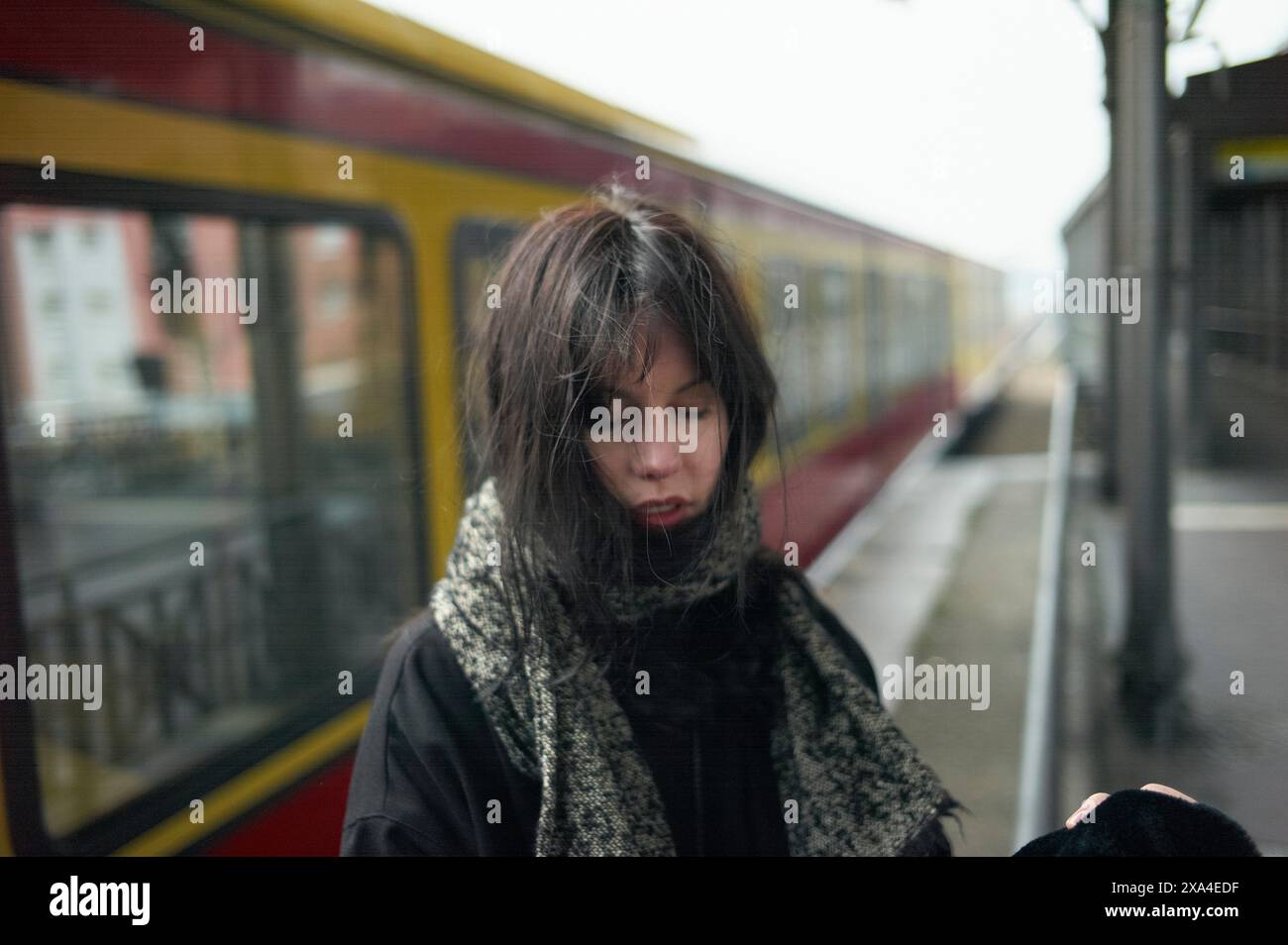 A woman stands on a train platform, her face partially obscured by windswept hair, as she appears deep in thought. Stock Photo