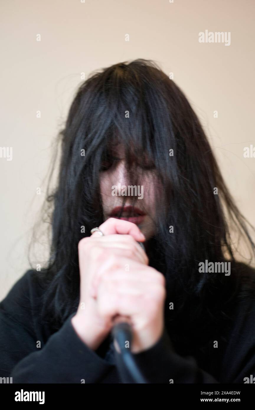A person with long black hair obscuring their face is holding a microphone close to their lips, set against a plain background. Stock Photo