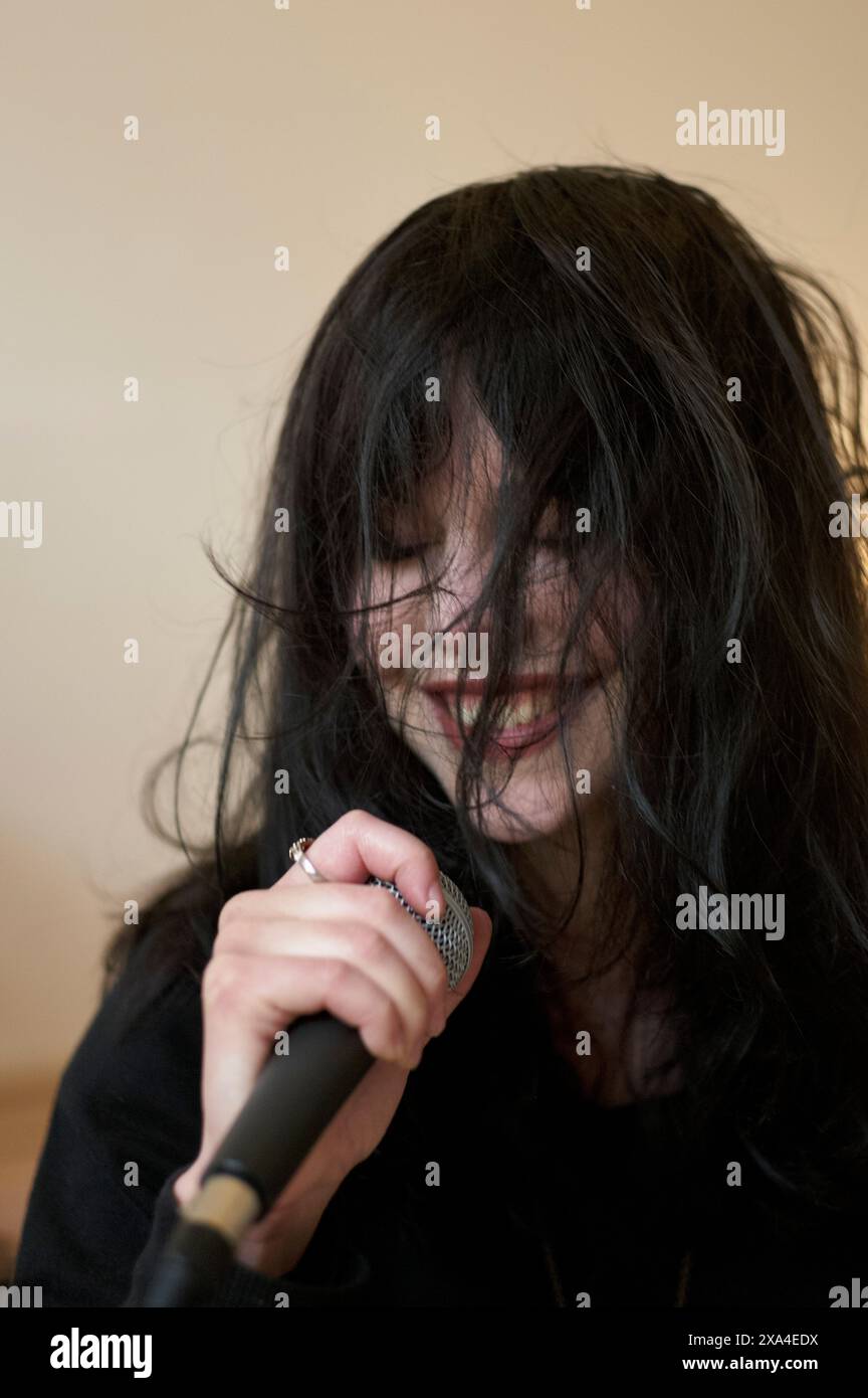 A person with long black hair is singing into a microphone with their head tilted down, eyes closed, and a joyful expression. Stock Photo