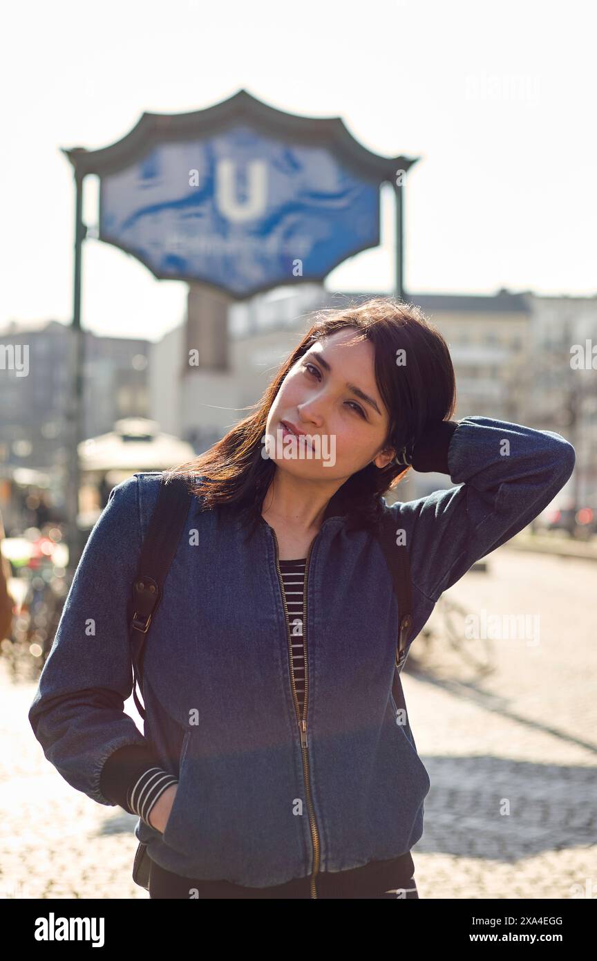 A woman stands outdoors with her hand in her hair, squinting slightly in the sunlight, with a blue U-Bahn sign in the background. Stock Photo