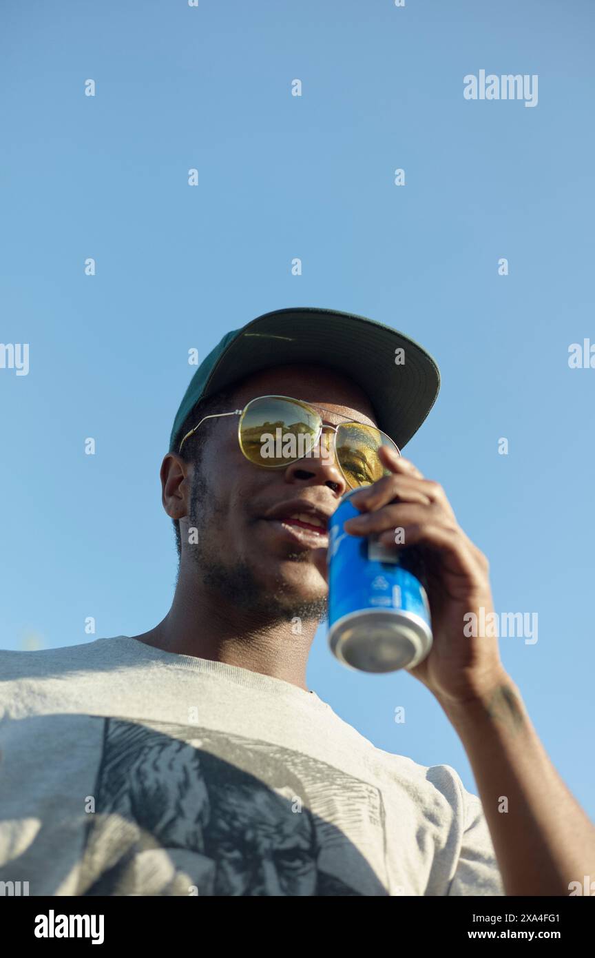 A man wearing sunglasses and a cap enjoys a beverage from a can against a clear sky. Stock Photo