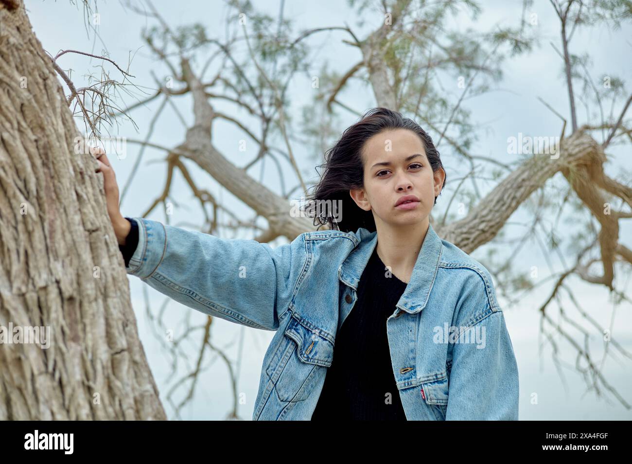 A person with shoulder-length hair stands outdoors, leaning casually against a tree, wearing a denim jacket over a black top, with a serious expression on their face and a barren tree and cloudy sky in the background. Stock Photo