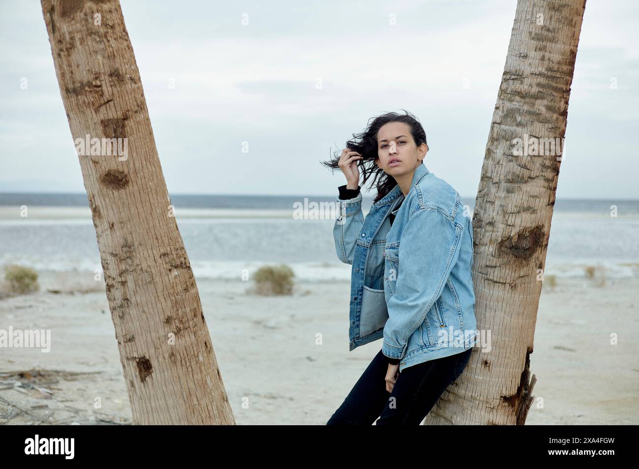 A person stands between two palm trees on a sandy beach, wearing a denim jacket and black pants, looking into the distance with a thoughtful expression. Stock Photo