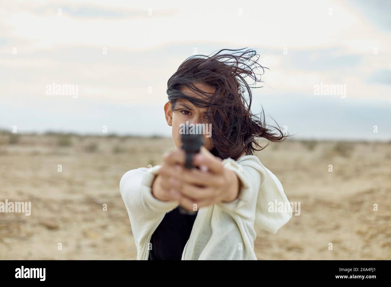 A person is pointing a gun directly at the camera with intense focus. Their hair is windswept, and they are dressed in a white jacket over a black top, set against a desolate landscape. Stock Photo