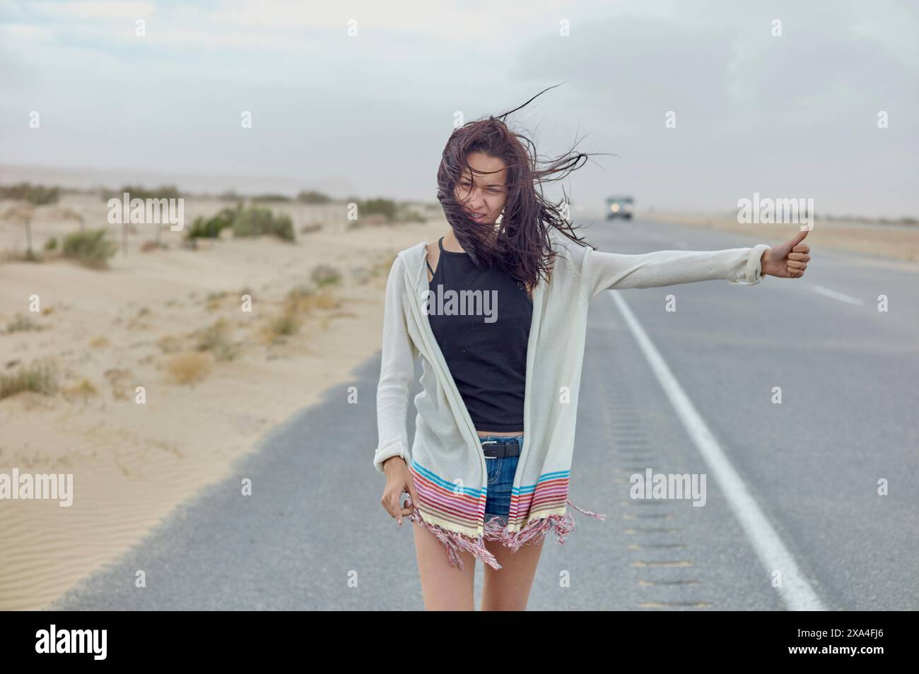 A young woman stands by a desert road, her hair tousled by the wind, as she hitches a ride with her thumb extended. Stock Photo
