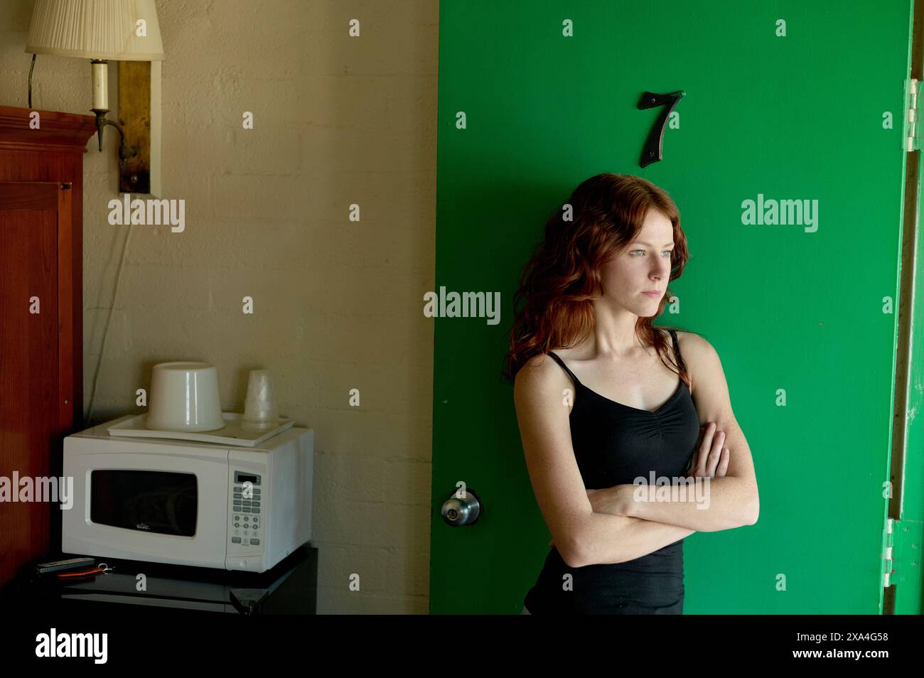 A woman stands in a room with her arms crossed, next to a kitchen counter with a microwave and a coffee maker, looking to the side against a green backdrop. Stock Photo