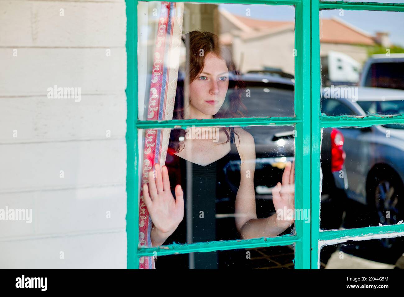 A woman is looking through a windowpane with her hands pressed against the glass, her expression one of contemplation or waiting. Stock Photo