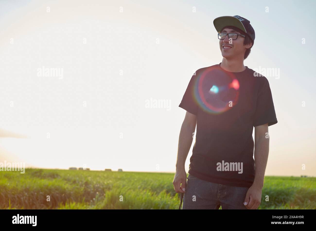 A smiling young man wearing glasses, a cap, and a black t-shirt stands in a field with the sun setting in the background, casting a warm glow. Stock Photo