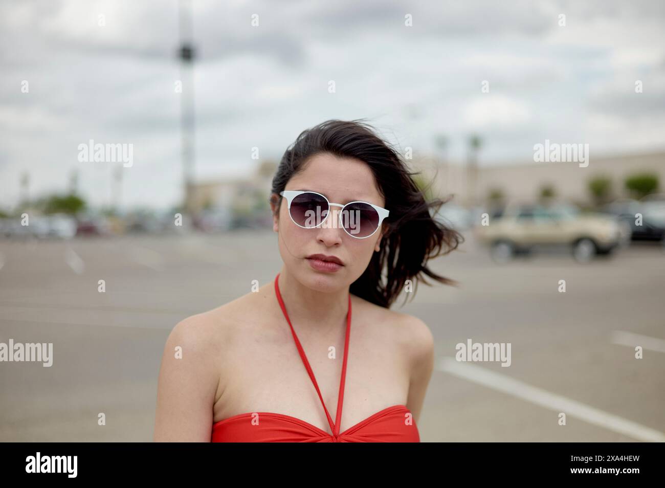 A woman wearing sunglasses and a red dress stands in a parking lot with a cloudy sky overhead and a classic car in the background. Stock Photo