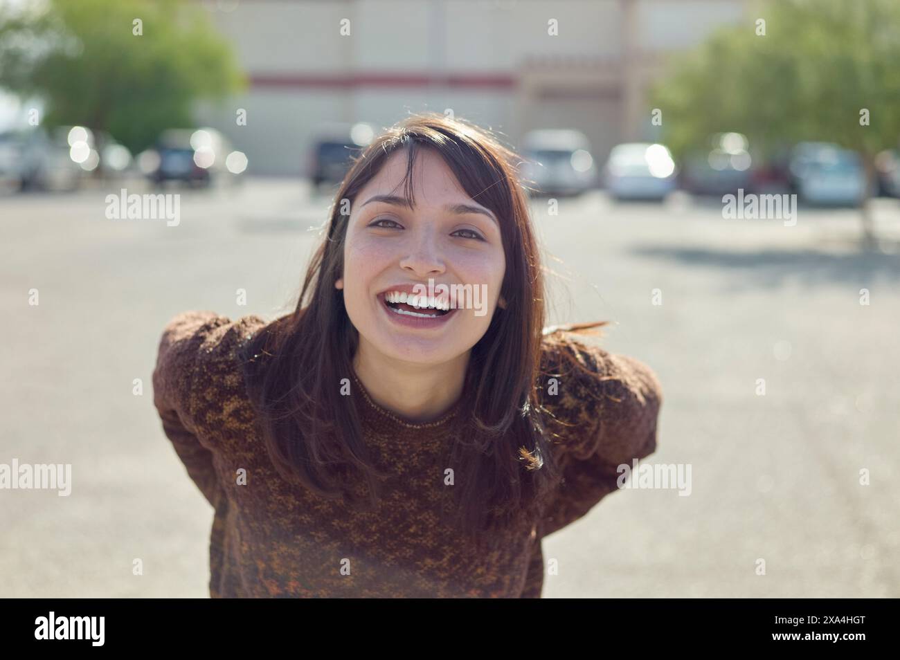 A young woman is smiling broadly with her arms outstretched, standing in a sunny parking lot with cars and a building in the background. Stock Photo