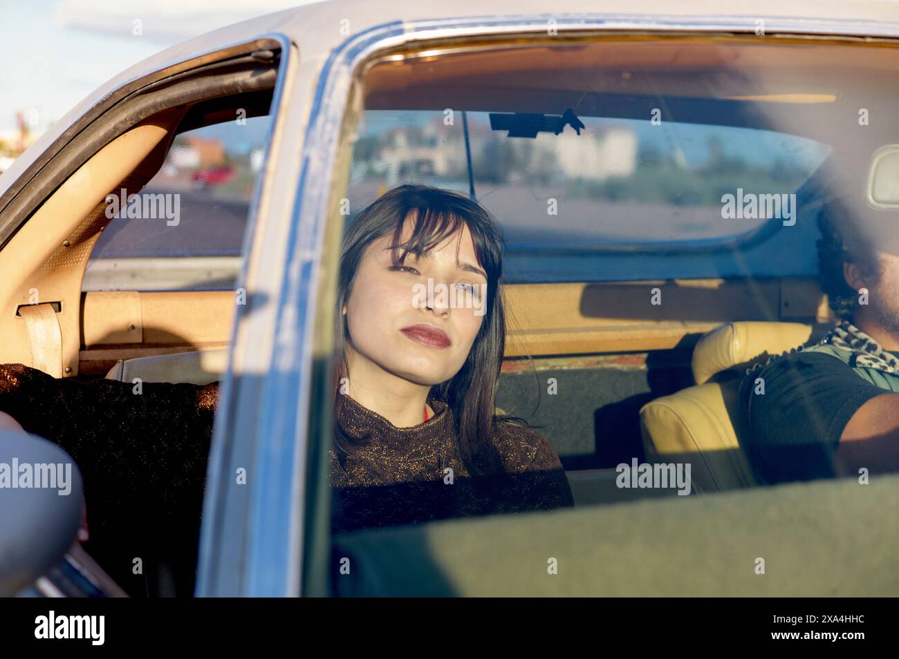 A woman with dark hair is seated in the front passenger seat of a vintage car, gazing out of the window with a thoughtful expression as the driver is partially visible with focus on the woman. Stock Photo