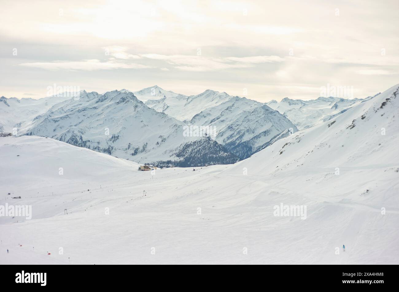 A serene winter landscape showcases snow-covered mountains with distant skiers and a ski lift cutting across the slope, under a soft cloudy sky. Stock Photo