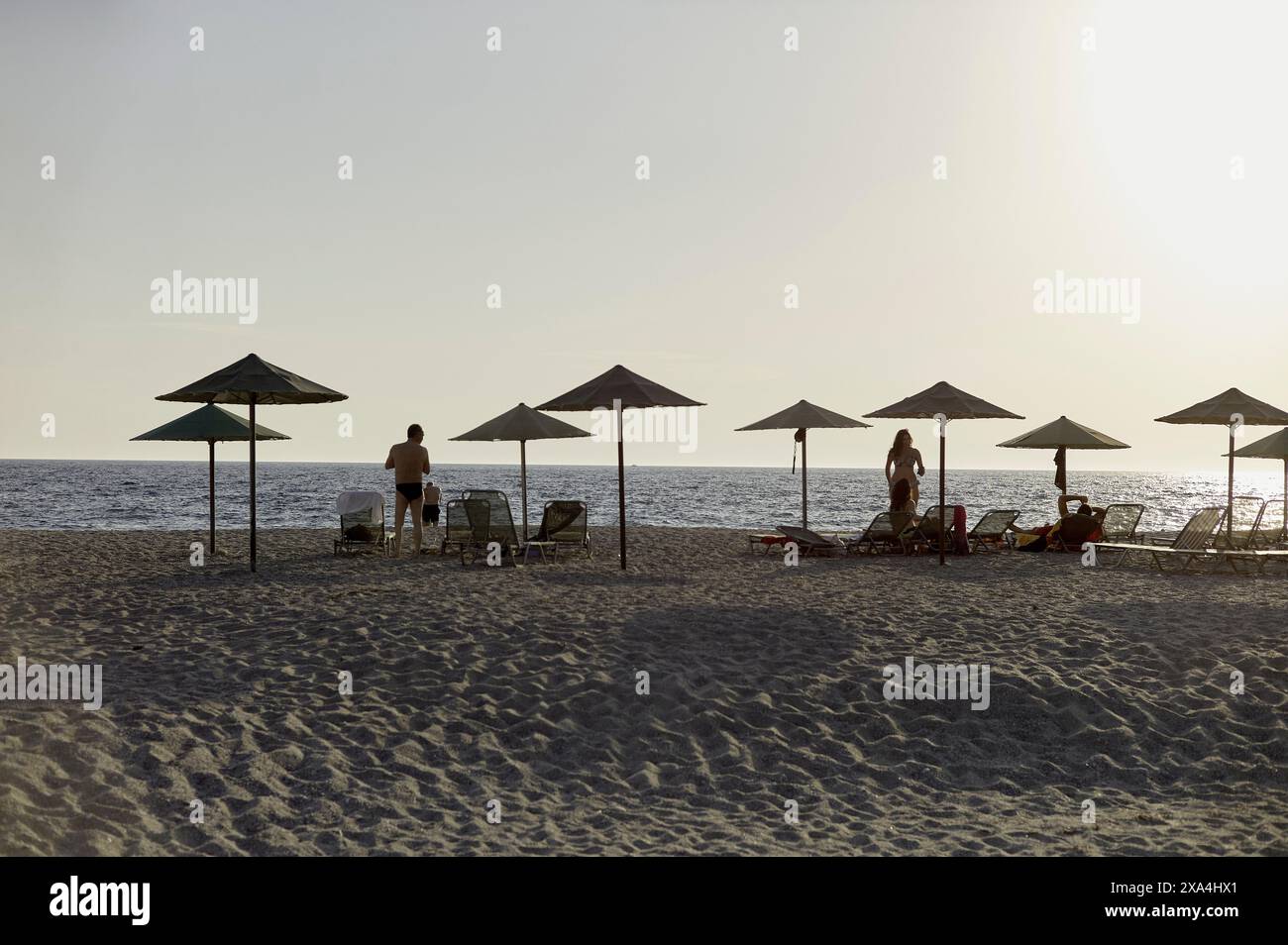 The image depicts a serene beach scene at sunset with several beach umbrellas, lounge chairs, and a few people enjoying the tranquil atmosphere. Stock Photo