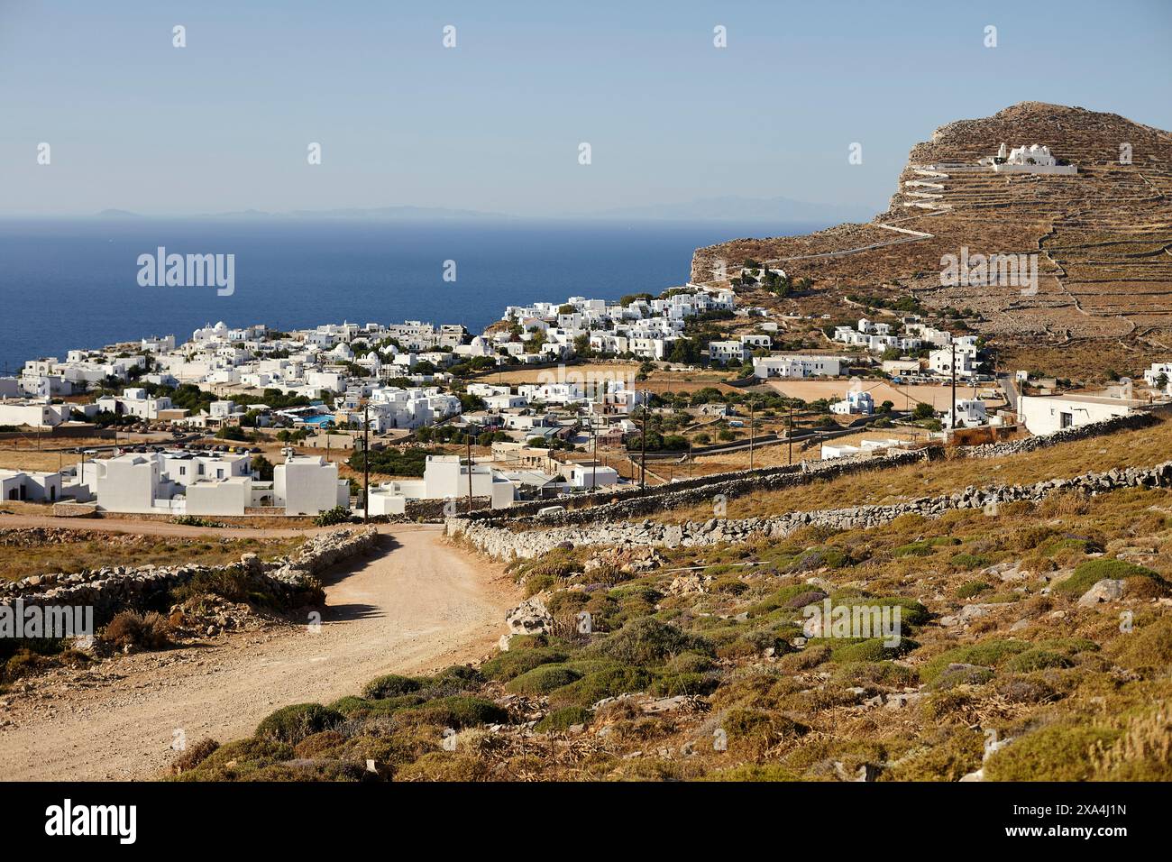 A scenic view of a coastal village with white buildings against a backdrop of clear blue skies, with a dirt path leading through a hilly landscape. Stock Photo