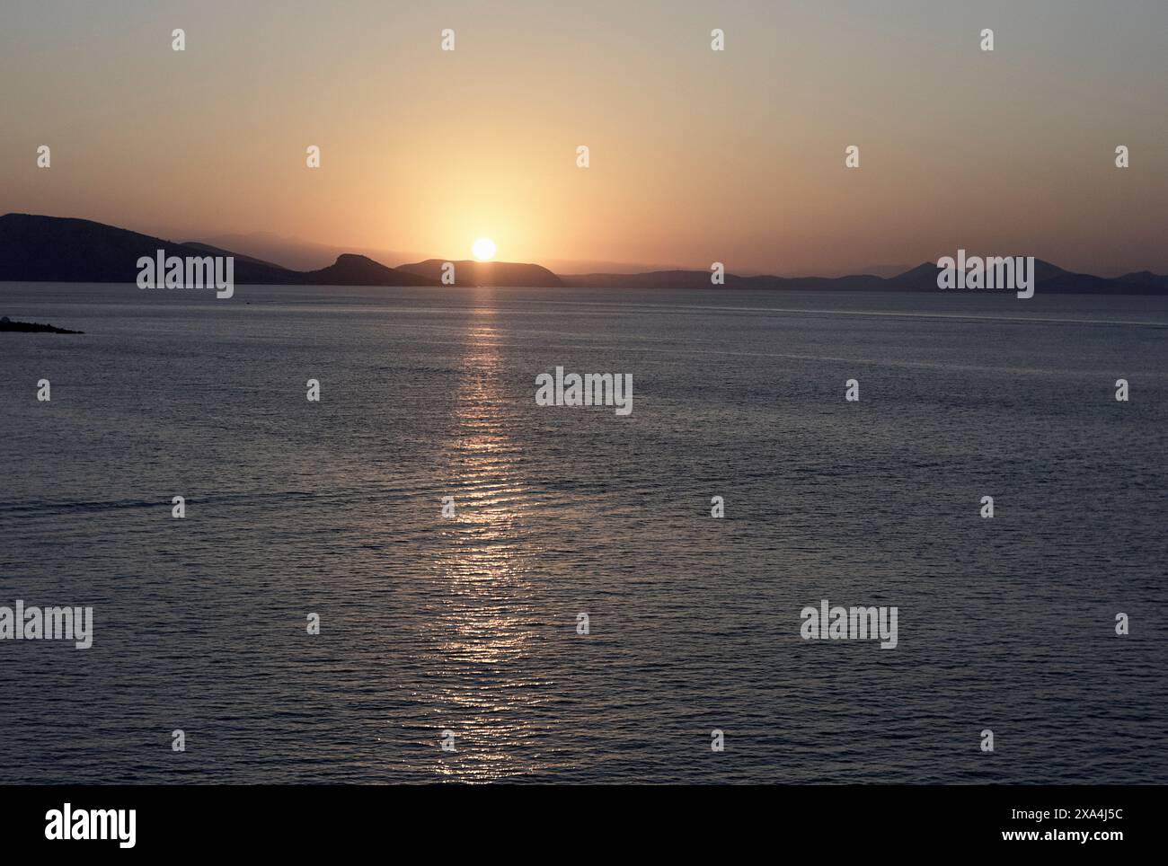 A serene sunset over the ocean with the sun casting a warm glow and a reflective path on the water, framed by distant silhouettes of mountains. Stock Photo