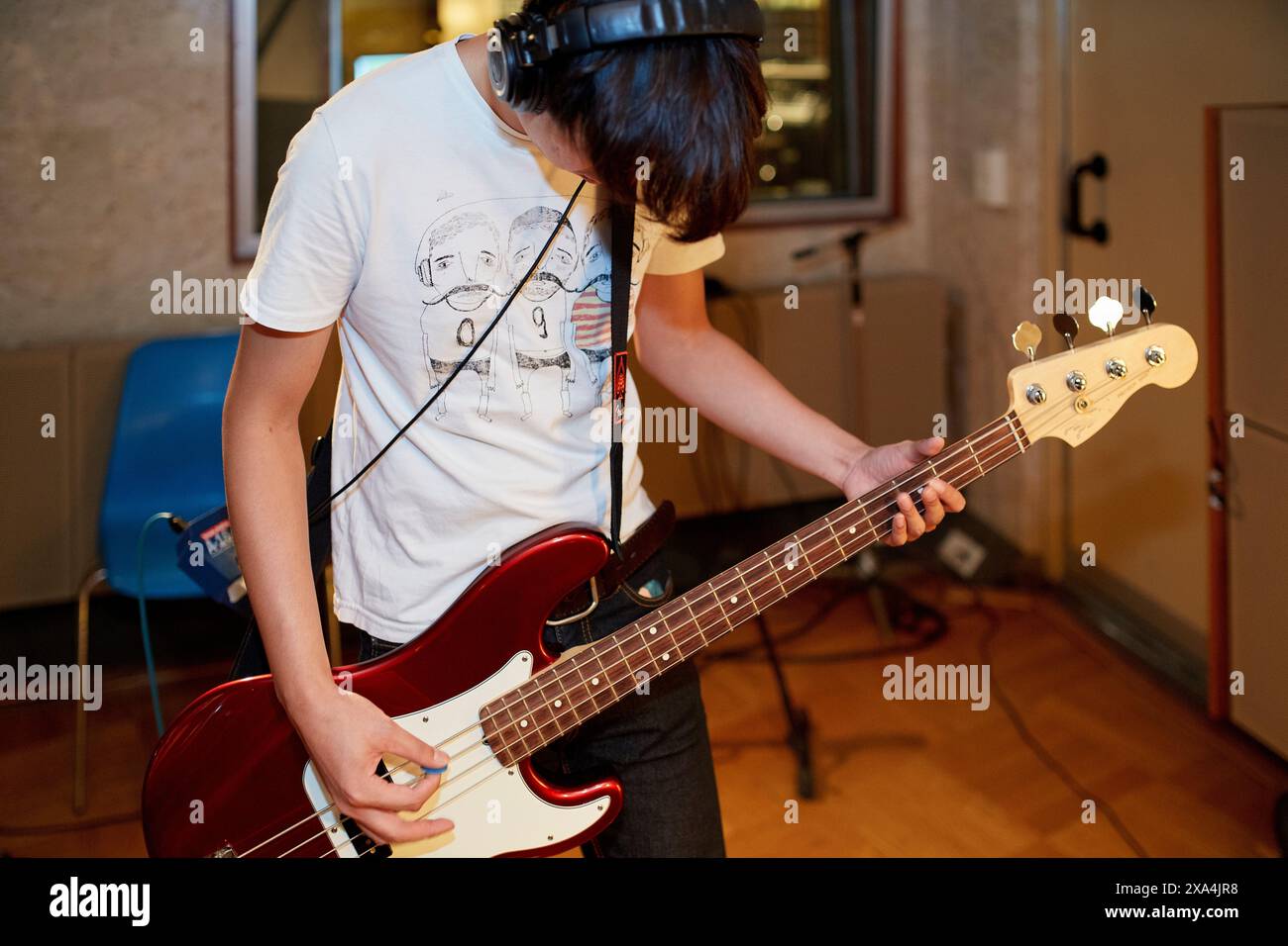 A person is engrossed in playing a red bass guitar while wearing headphones in a recording studio, with musical equipment in the background. Stock Photo