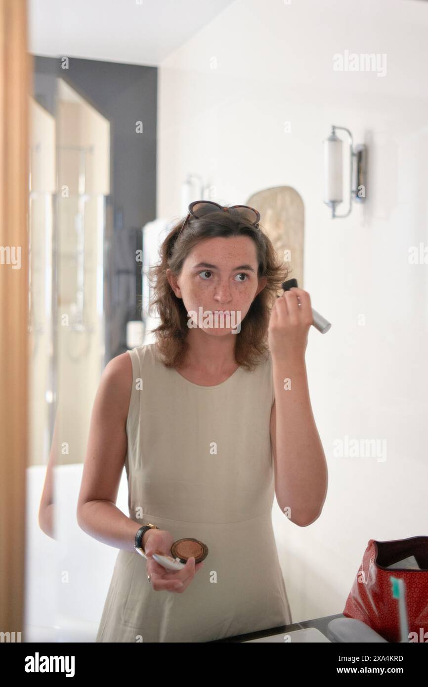 A woman is standing in front of a mirror holding a perfume bottle, preparing to spray it. She appears focused, with a neutral expression. Stock Photo