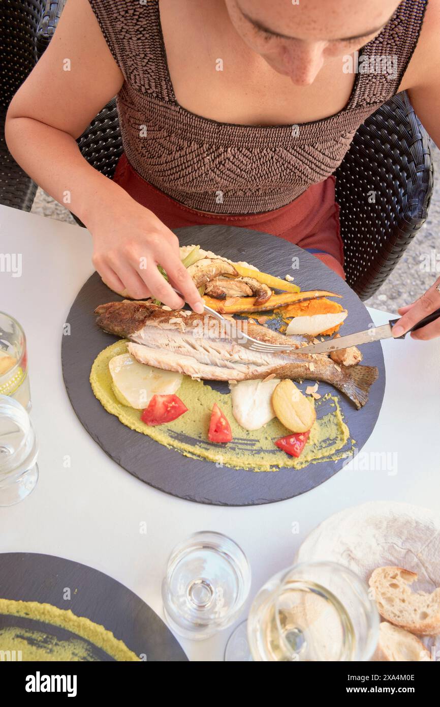 A person is seated at a table, focused on slicing a grilled fish served with slices of lemon, herbs, and vegetables on a round slate plate. Stock Photo