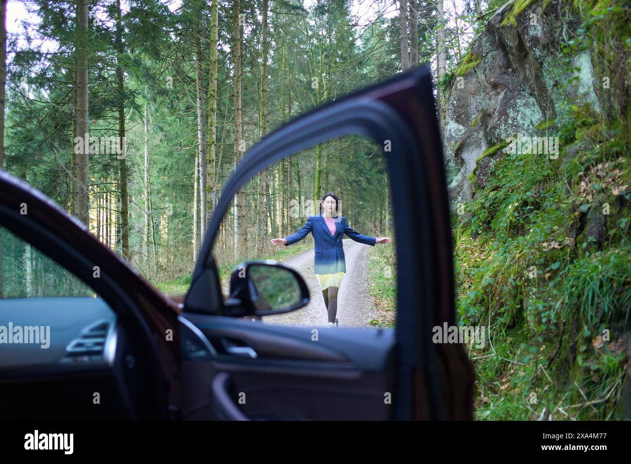 A woman is standing in the middle of a forest road, viewed through an open car door from the driver's seat perspective. Stock Photo