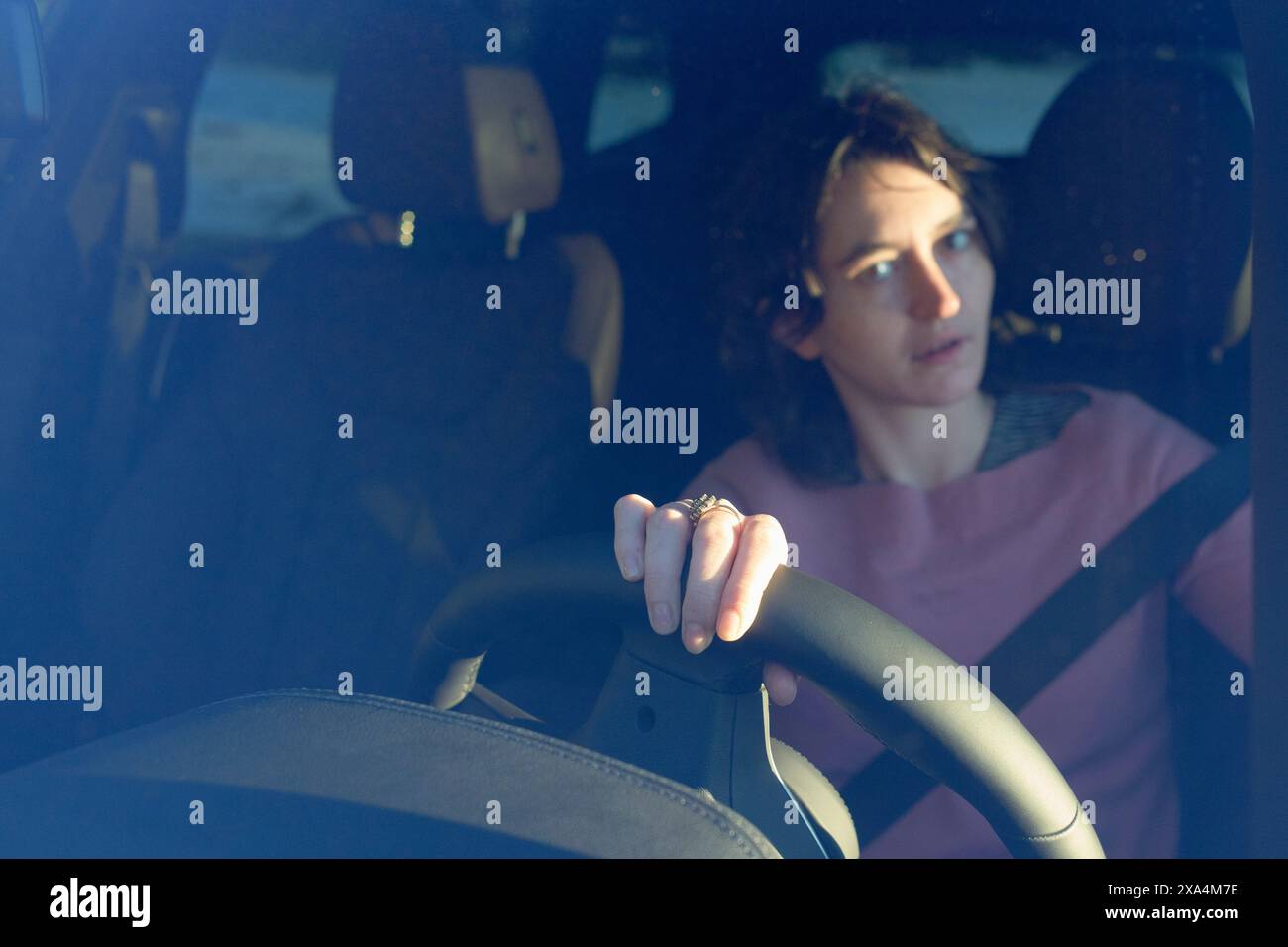 A woman is sitting behind the steering wheel in a vehicle, looking pensive as she gazes out of the driver's side window, with the sun casting shadows across the interior. Stock Photo