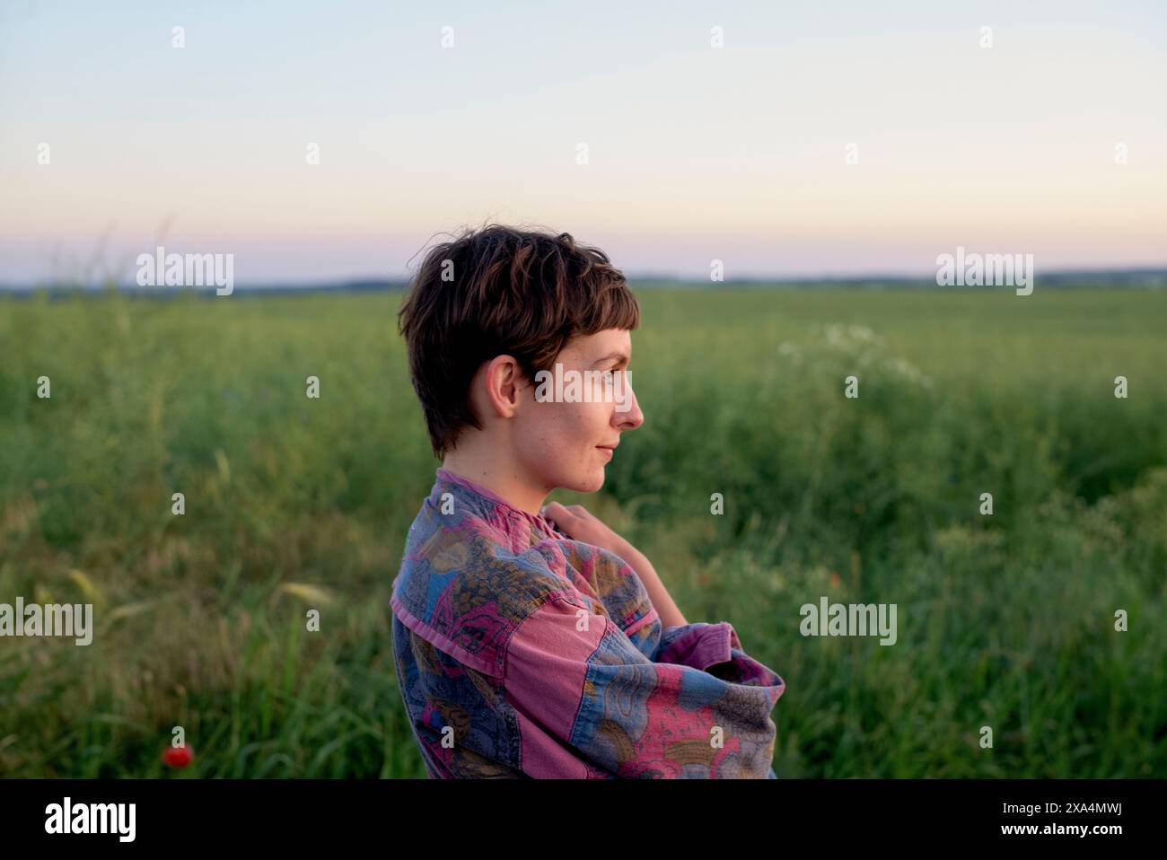 A young individual with short hair sits in a field at dusk, gazing into the distance with a serene expression. Stock Photo