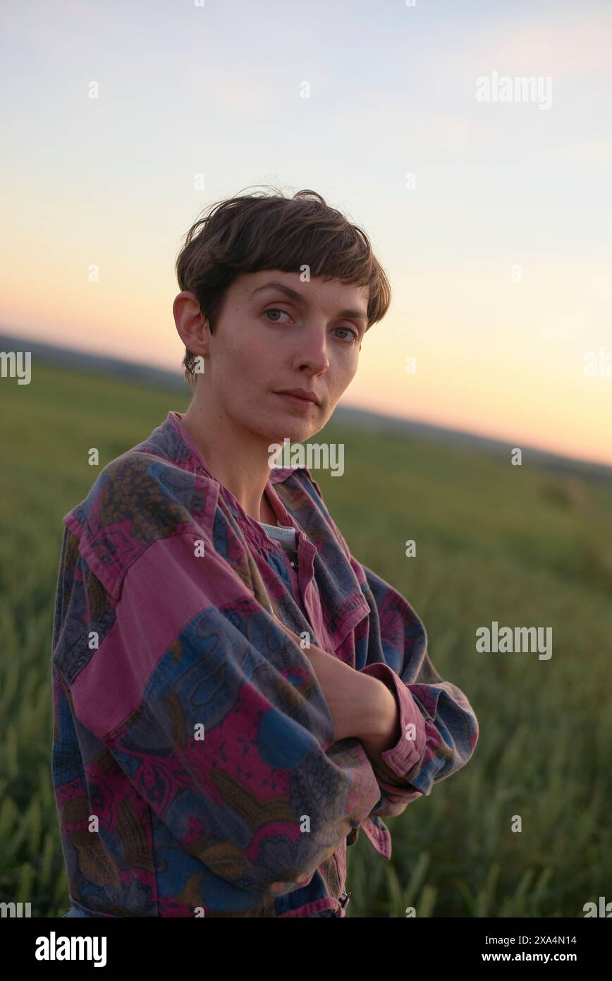 A person with short hair stands in a field at dusk, arms crossed and looking at the camera with a serene expression, wearing a colorful patterned shirt. Stock Photo
