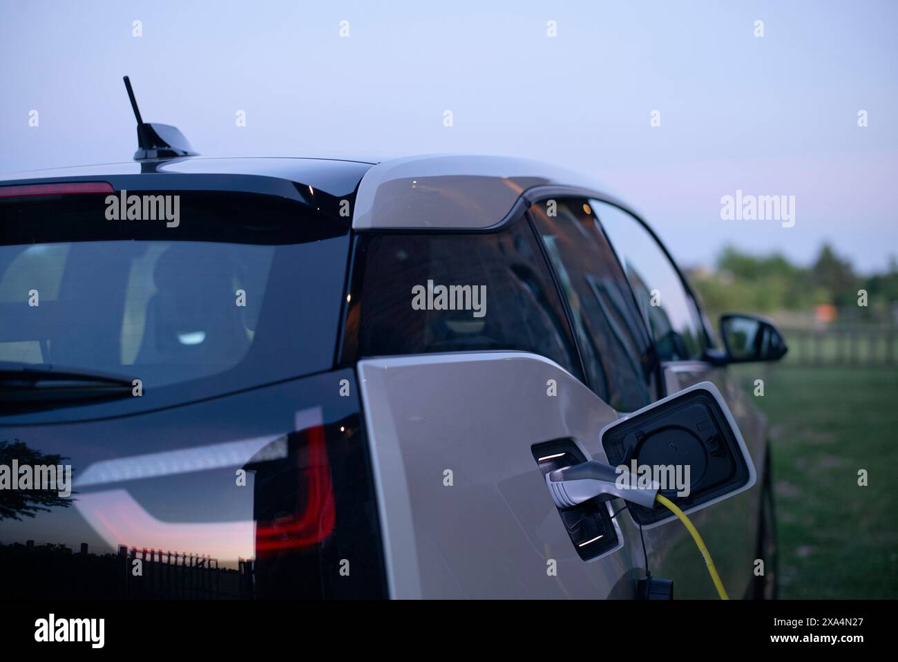An electric car is charging at a station during twilight, with the charge cable connected to the port on the vehicle's side. Stock Photo