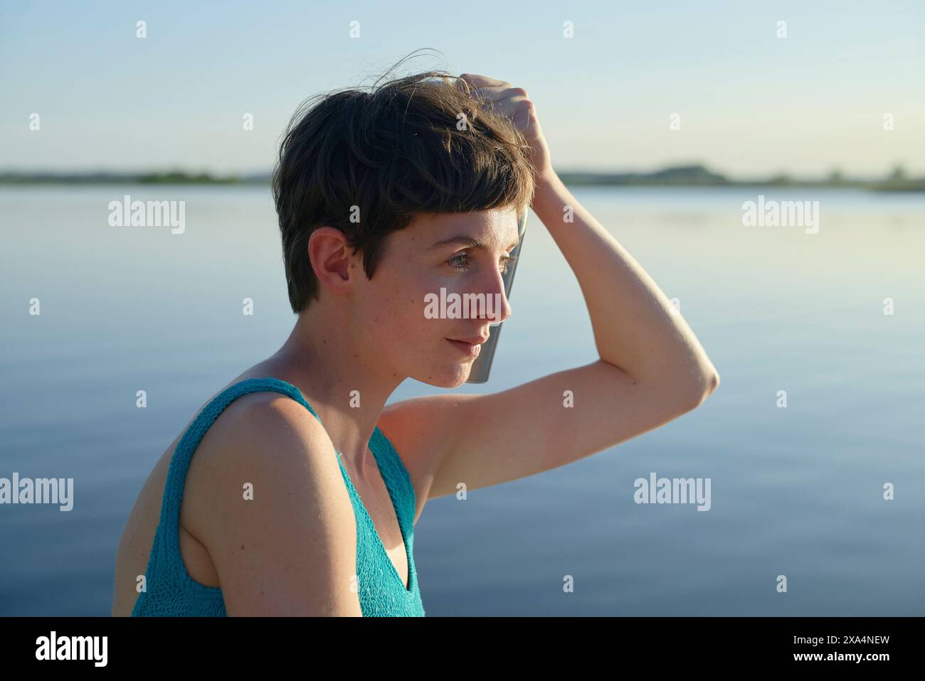 A young woman with short hair stands by a body of water at dusk, lifting their hand to their head, with a thoughtful expression and the horizon in the background. Stock Photo