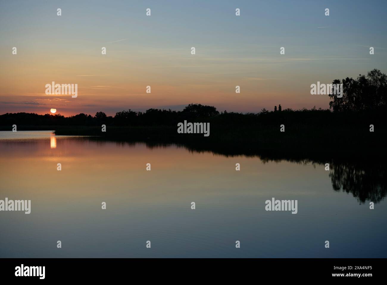 A serene sunset scene over a calm lake with reflections of the diminishing sun and silhouetted landscape against a gradient evening sky. Stock Photo