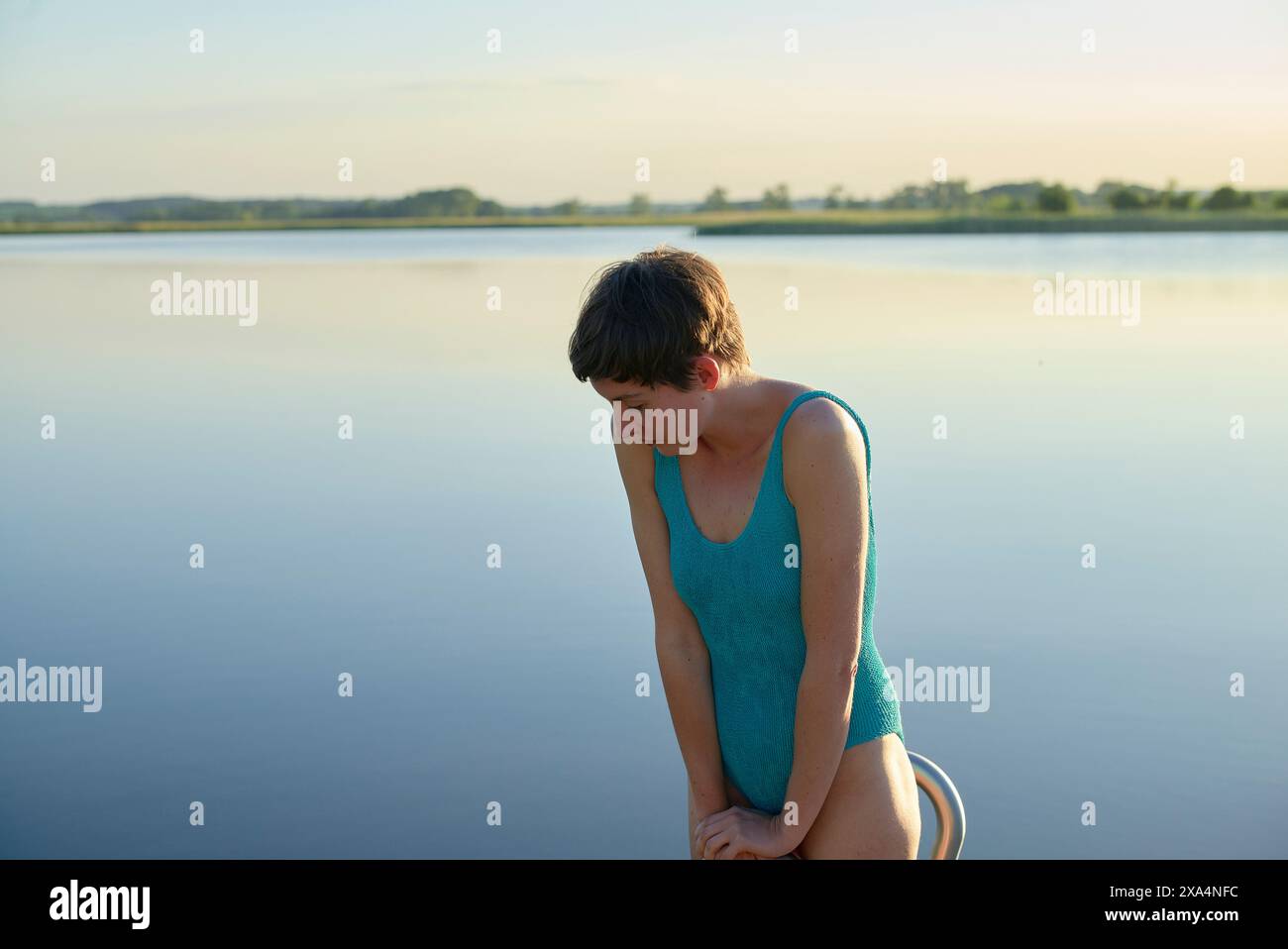 A young woman in a blue swimsuit sits contemplatively by a calm lake ...