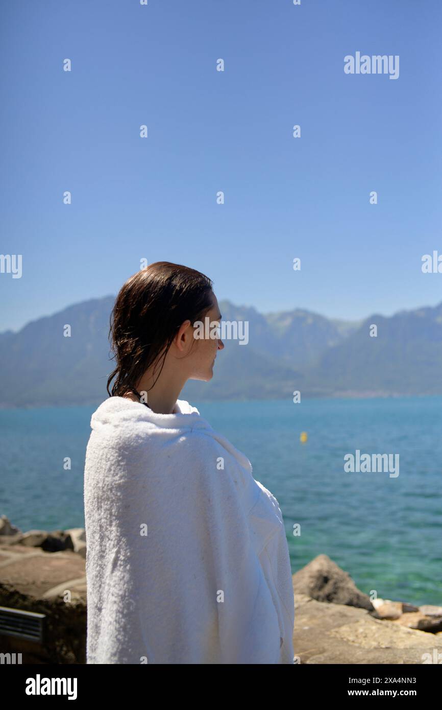 A person wrapped in a white towel stands by a serene lakeside with mountains in the background, gazing at the picturesque view. Stock Photo