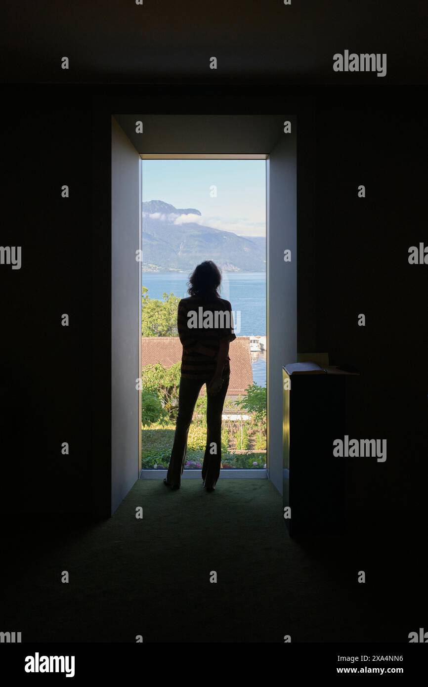 A silhouette of a person standing at an open doorway looking out towards a panoramic view of a serene lake and mountains under clear, sunny skies. Stock Photo