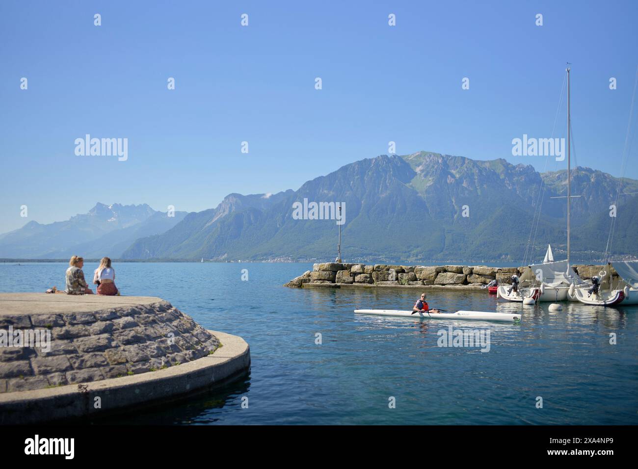 A scenic lakeside view where two people are seated on the edge of a pier, looking out at the water where a person is kayaking near docked sailboats, with mountains in the background under a clear blue sky. Stock Photo
