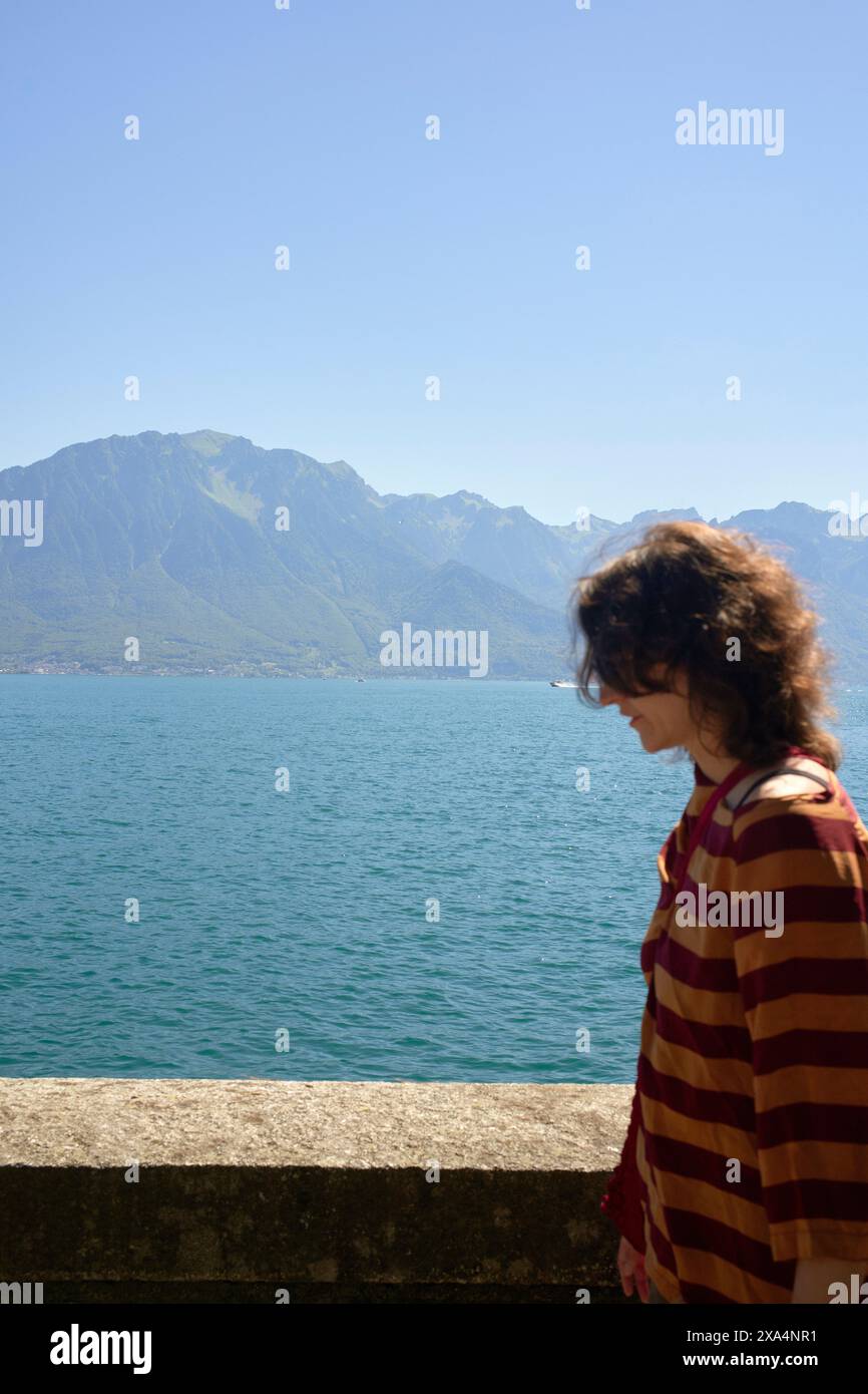 A person stands in profile with a serene lake and mountain landscape in the background underneath a clear blue sky. Stock Photo