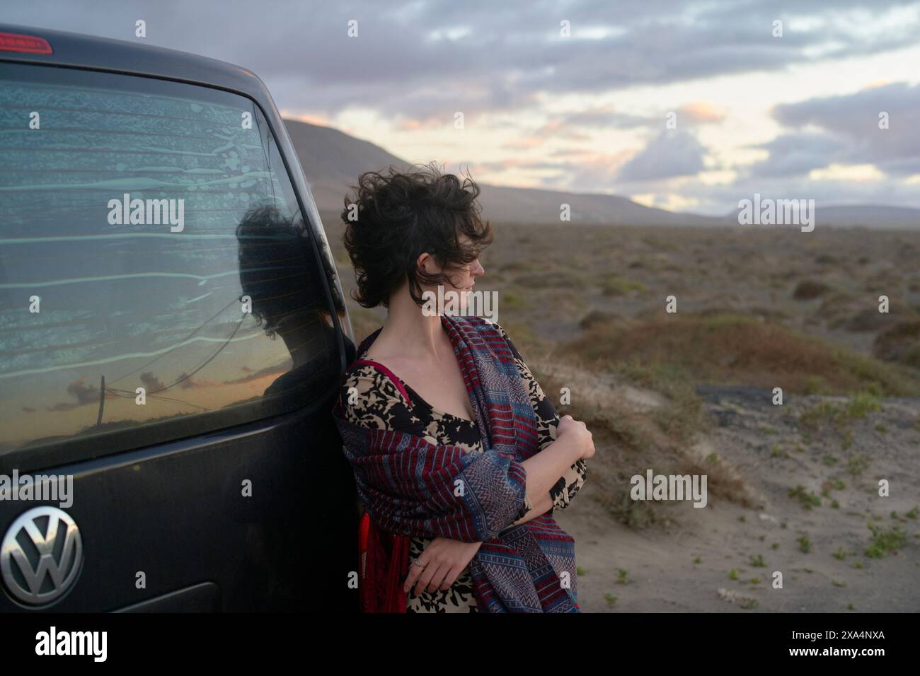 A woman leans against a Volkswagen vehicle with a cracked windshield, looking into the distance at a desolate landscape under a cloudy sunset sky. Stock Photo