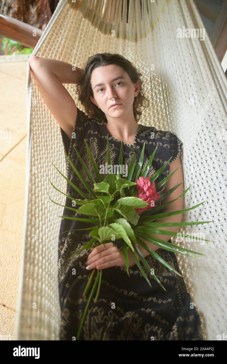 A young woman lounges in a white hammock holding a bouquet of green leaves and a red flower, dressed in a black lace garment, with a relaxed and contemplative expression. Stock Photo