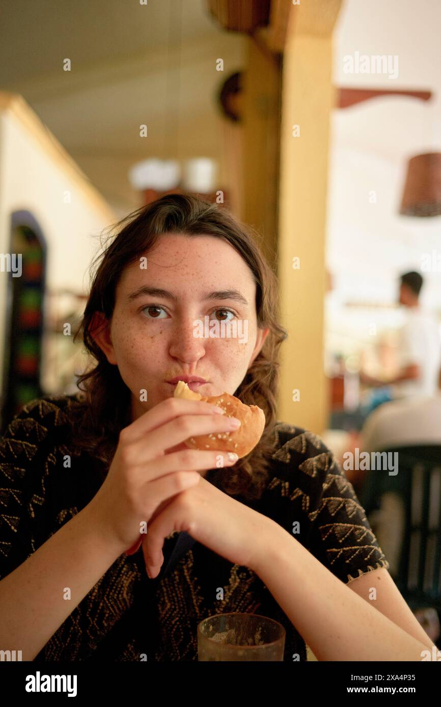 A woman is sitting at a table eating a sandwich with a thoughtful expression, in a casually-styled indoor setting with a background that suggests a restaurant or cafe ambiance. Stock Photo
