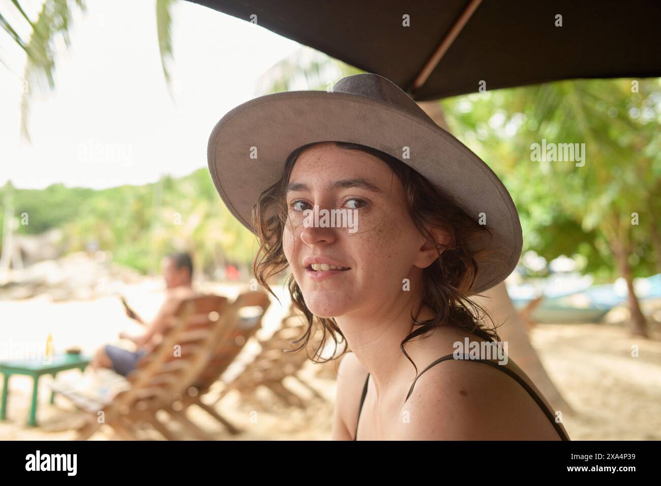 A woman wearing a gray hat and a black top sits under a parasol at a beach, with a sandy shore and trees in the soft-focused background. Stock Photo