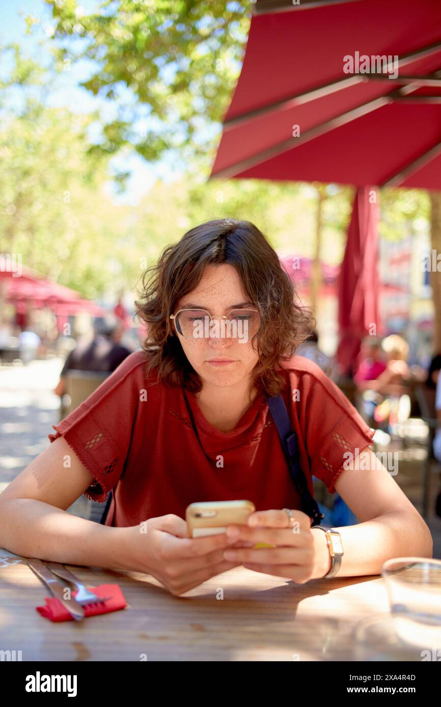 A young woman is sitting at a cafe table outdoors, focused on their smartphone, with sunlight filtering through the trees. Stock Photo