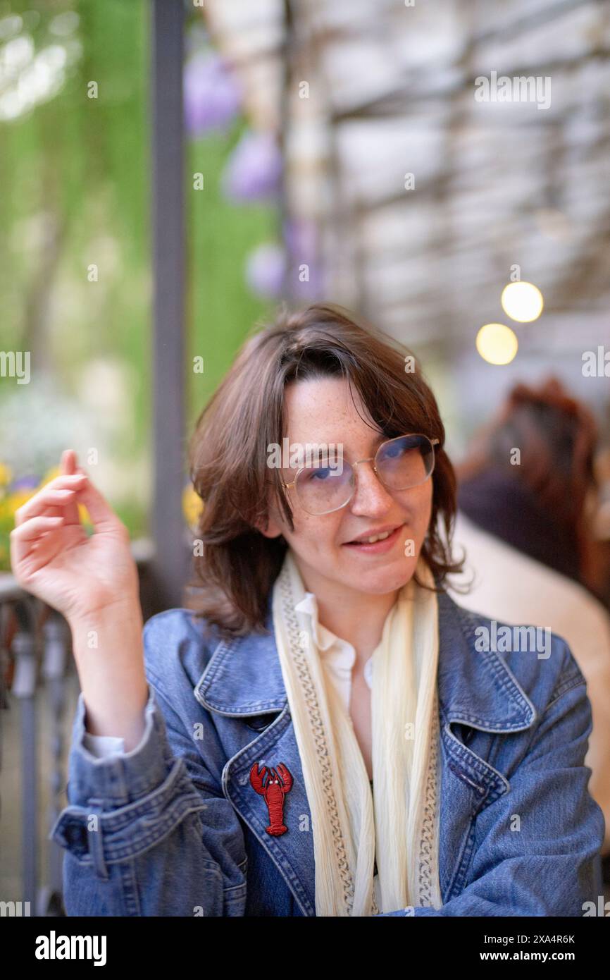 A smiling woman sits at an outdoor cafe, holding a yellow flower, wearing a denim jacket, glasses, and a white scarf. Stock Photo