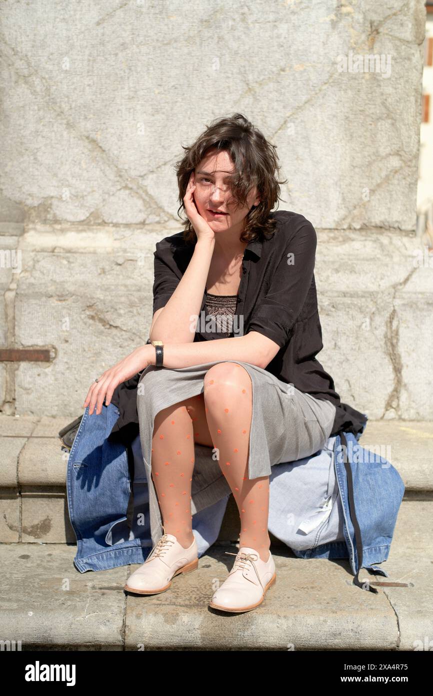 A woman is sitting on steps outside, smiling with her chin resting on her hand, wearing a black jacket, grey dress, and patterned tights, with a denim jacket placed beside her. Stock Photo
