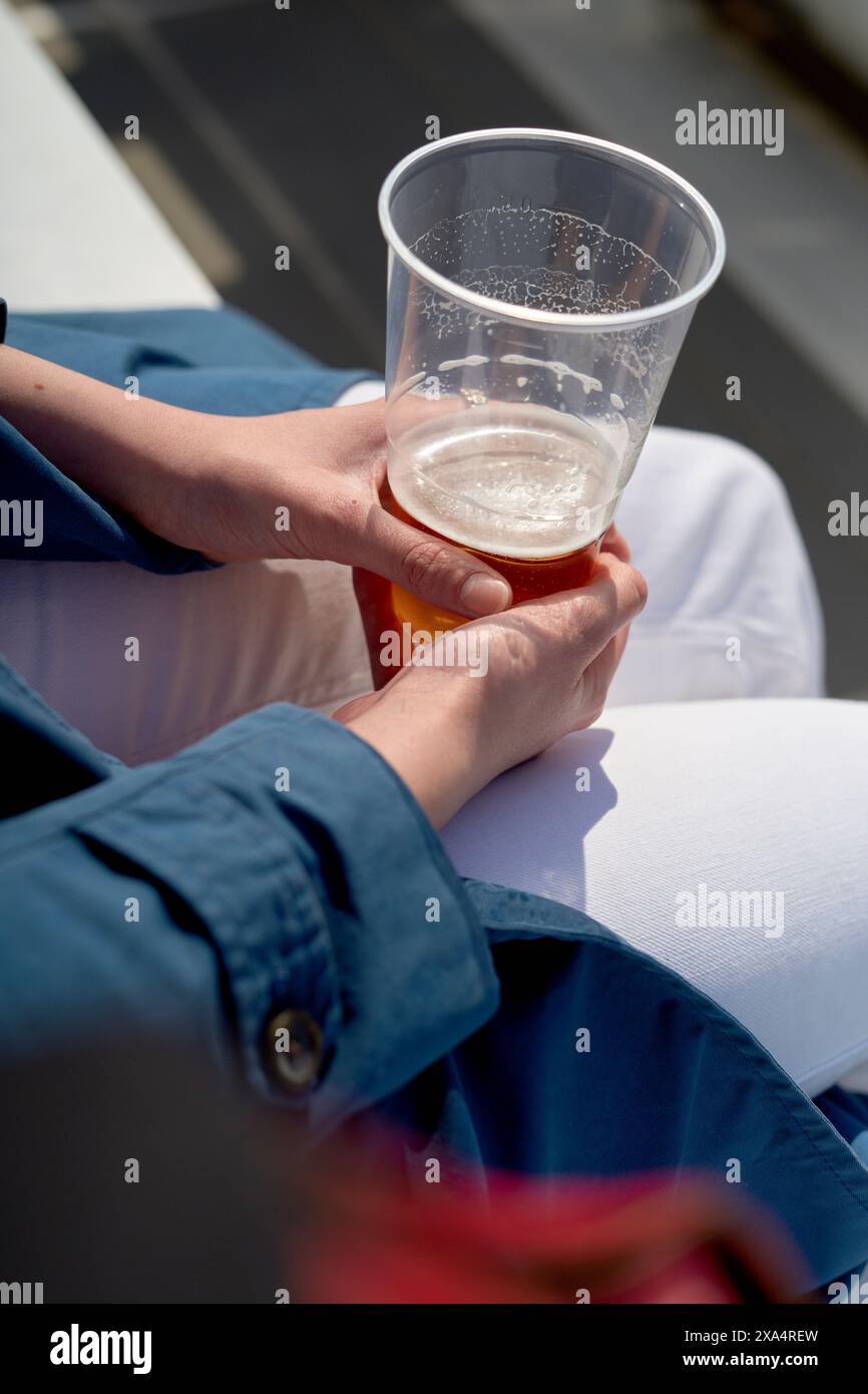 Sunlit hands cradling a half-full plastic beer cup, all set against a casual outdoor backdrop. Stock Photo