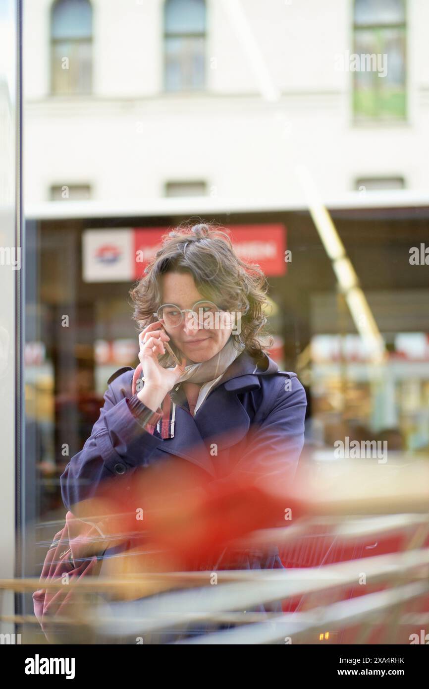 Woman in blue coat talking on the phone behind a blurred red object in the city. Stock Photo