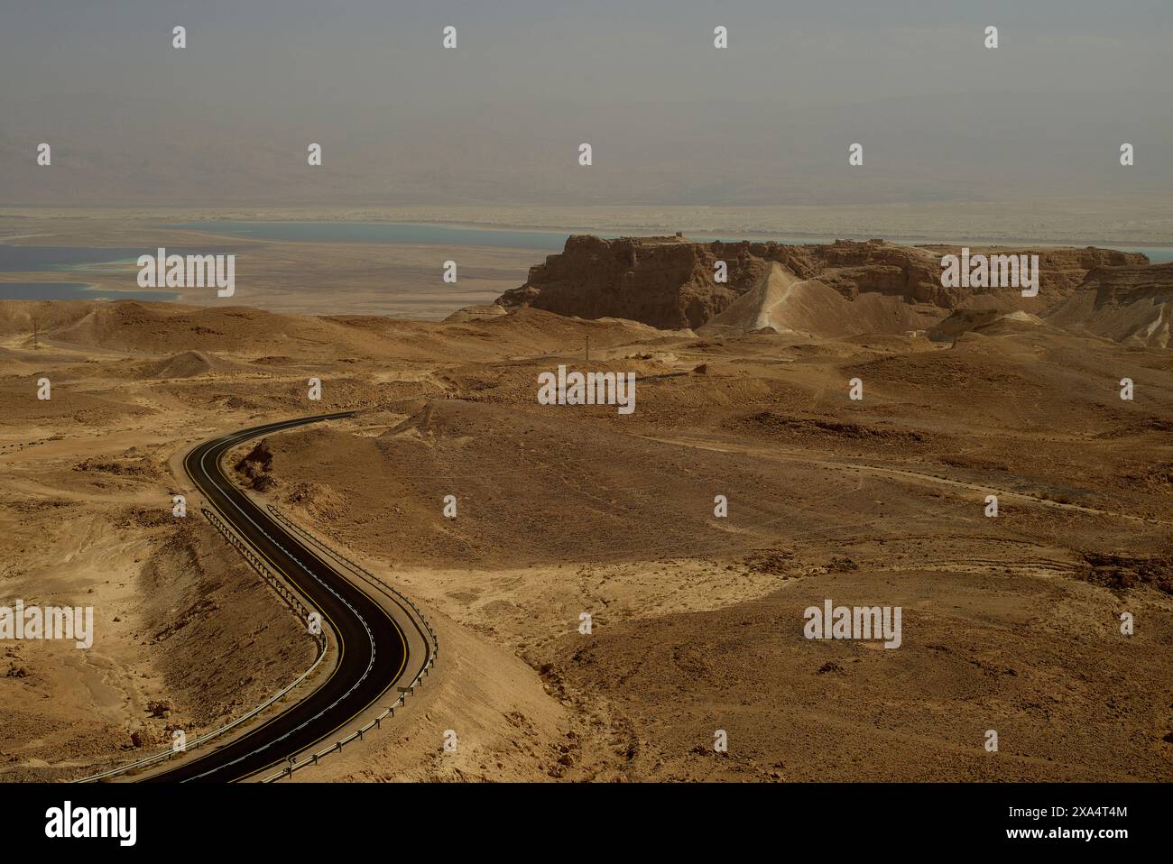 Winding road cutting through a desert landscape with a hint of an oasis in the distance under a hazy sky. Stock Photo