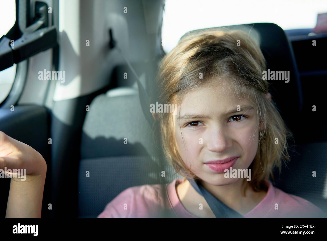 Young girl smiling gently while seated in a car wearing a seatbelt, with sunlight filtering through the window. Stock Photo
