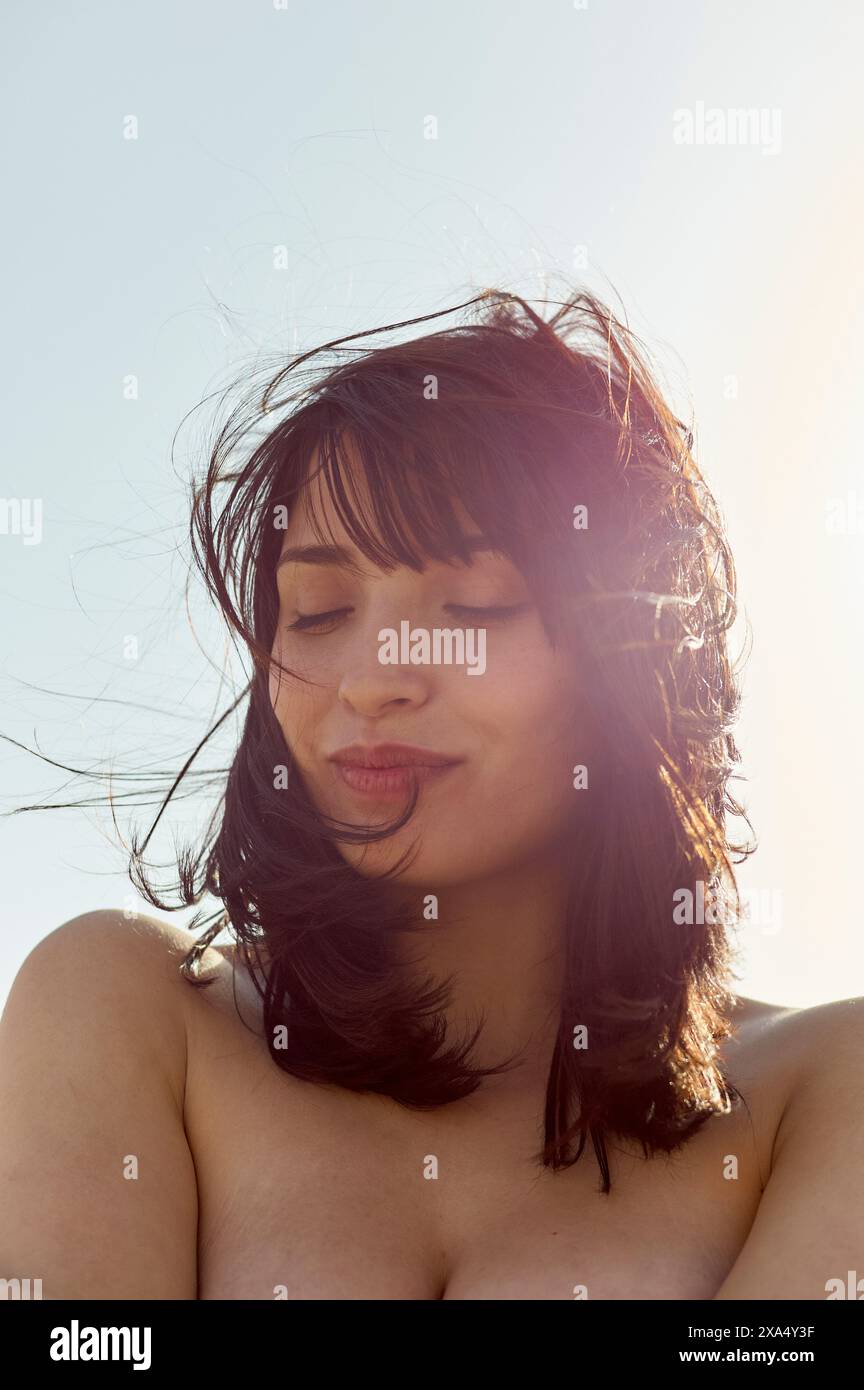 Sunlit portrait of a content woman with her eyes closed and hair tousled by the wind. Stock Photo