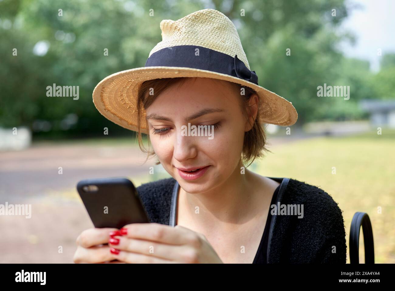 A smiling woman wearing a sun hat is looking at her smartphone in a park setting. Stock Photo